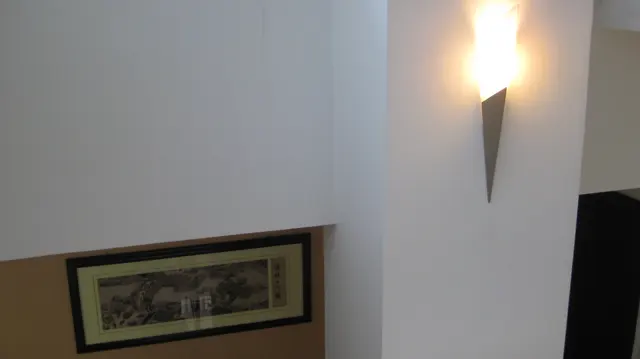 A glowing wall light hangs above and beside a framed picture on a pale interior wall.
