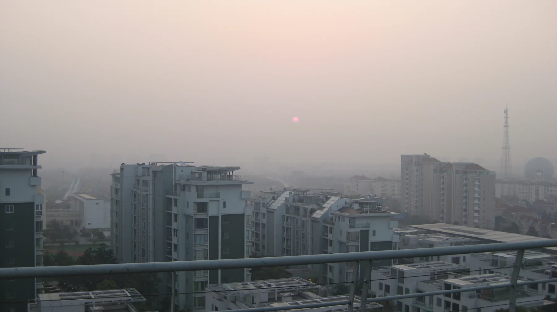 A faint sun hangs in a hazy peach-coloured sky above apartment blocks and a foreground balcony railing.