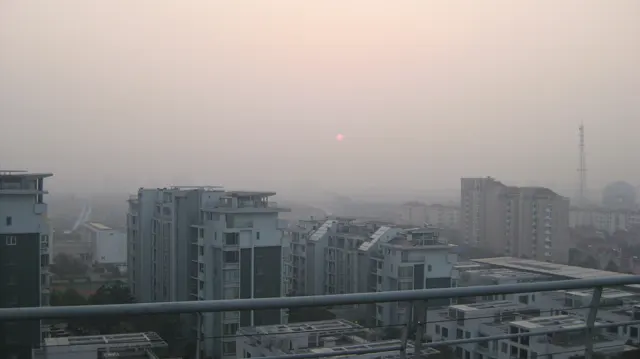 A faint sun hangs in a hazy peach-coloured sky above apartment blocks and a foreground balcony railing.