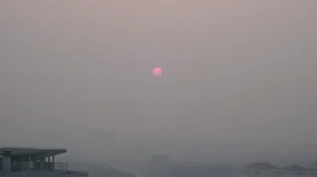 A small pink sun glows through a dense grey-pink haze above barely visible buildings.