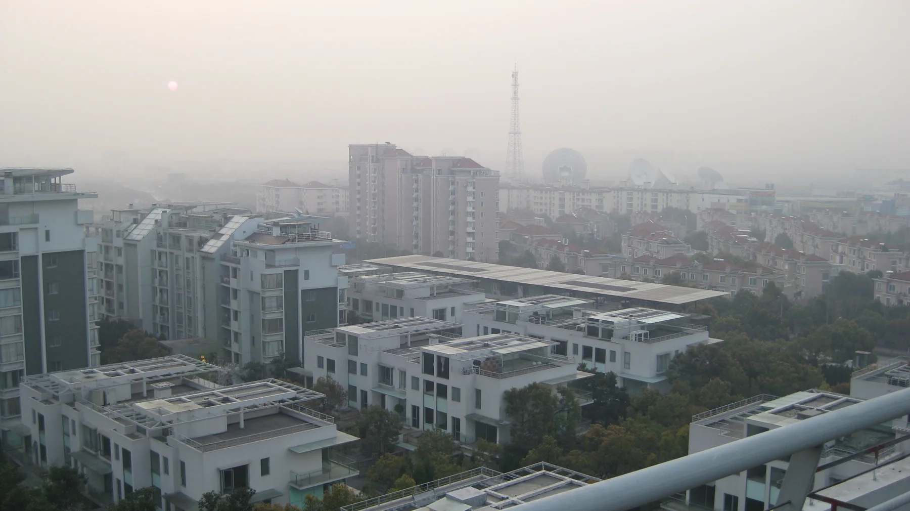 Flat-roofed residences lead towards distant apartment blocks and a slender tower beneath a pale sky.