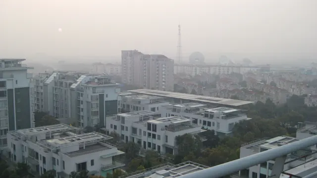 Flat-roofed residences lead towards distant apartment blocks and a slender tower beneath a pale sky.