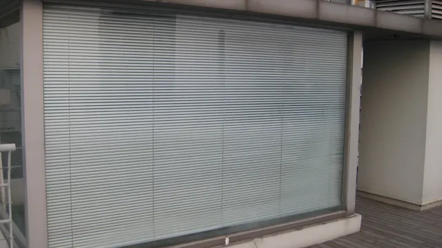 Closed horizontal blinds cover a broad glazed opening beside a narrow outdoor deck.