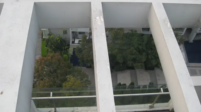 An overhead view through pale structural beams looks down towards a planted courtyard and balconies.