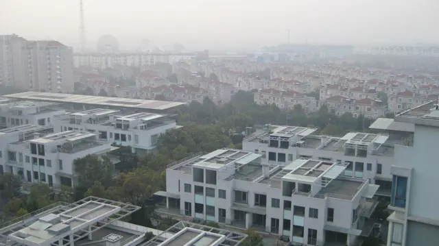 An elevated view takes in rows of white flat-roofed homes and a dense hazy city beyond.