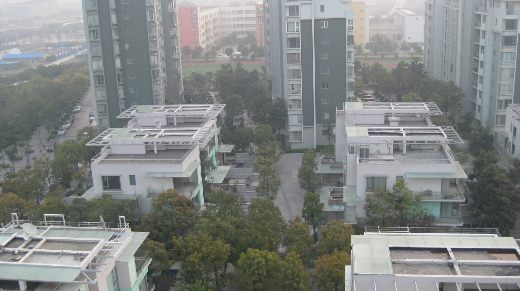 A straight landscaped path runs between white residences towards tall apartment blocks, seen from above.