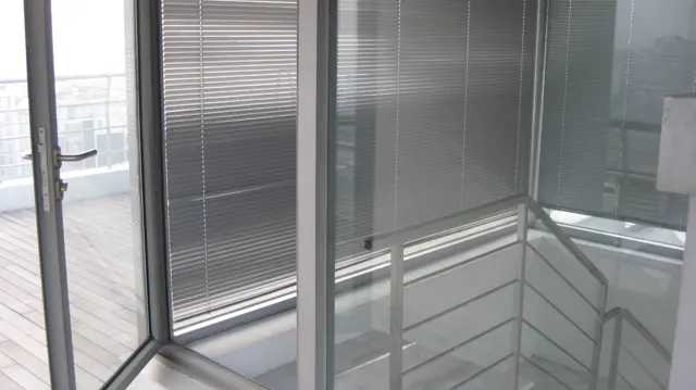 A stair landing with metal railings sits beside floor-to-ceiling glazing and horizontal blinds.