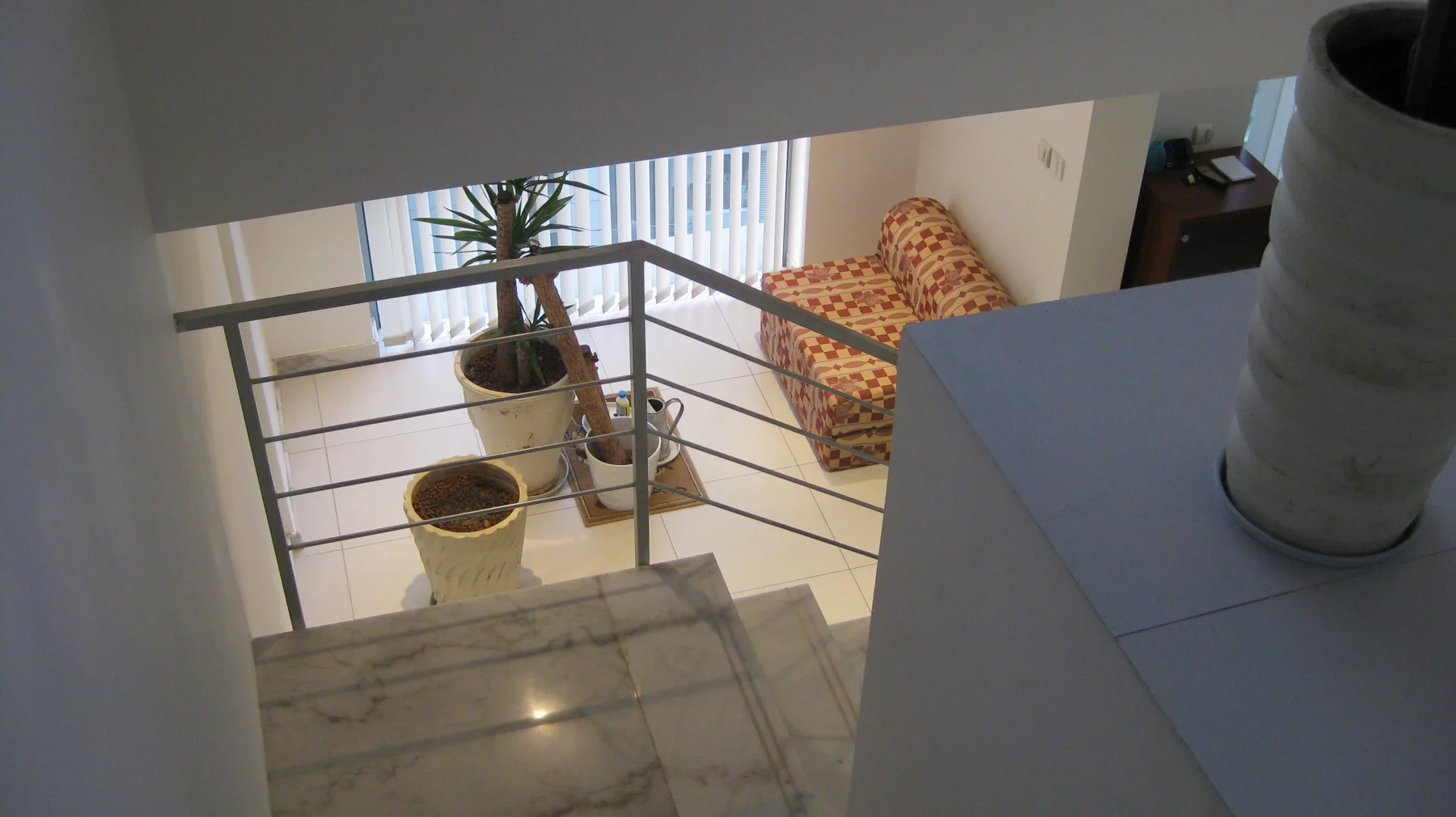 Pale steps descend beside a metal railing towards plants and a patterned seat on the lower floor.