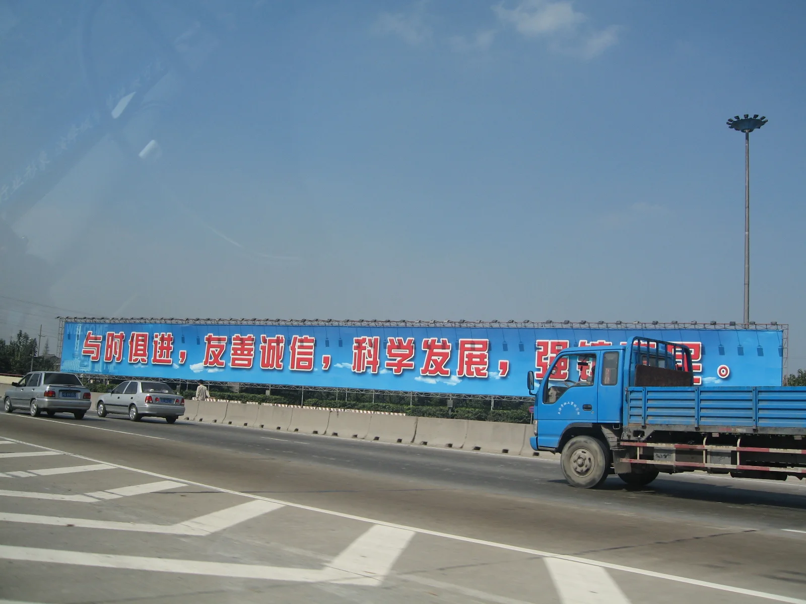 A blue truck passes a roadside construction hoarding carrying large Chinese lettering beneath a clear sky.