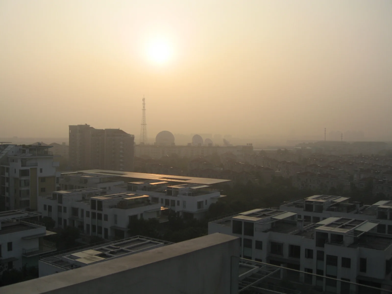A low sun shines through golden haze above a neighbourhood of flat-roofed buildings.