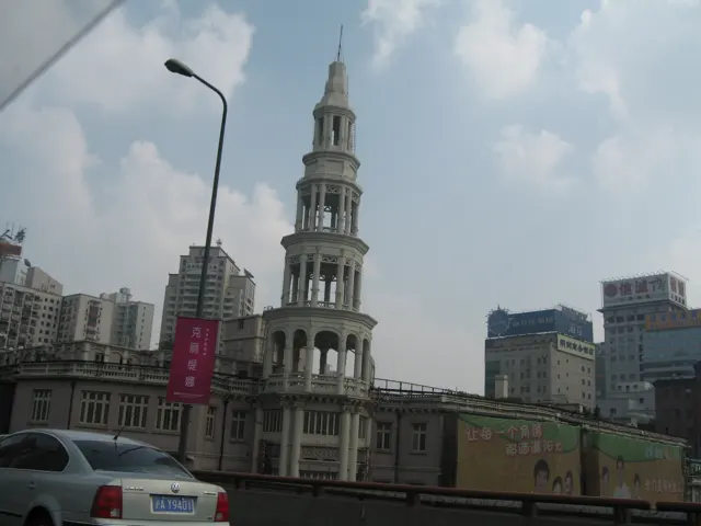 A pale tiered tower rises above a low building beside a road, with city blocks and clouds behind.