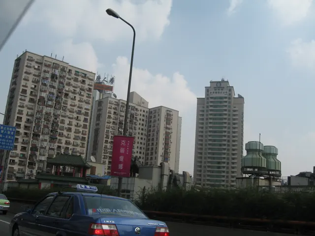 Cars pass a row of tall residential and commercial buildings beneath a street lamp and a partly cloudy sky.