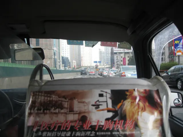 An interior view past a front seat and an advertisement looks through a vehicle windscreen towards city traffic.