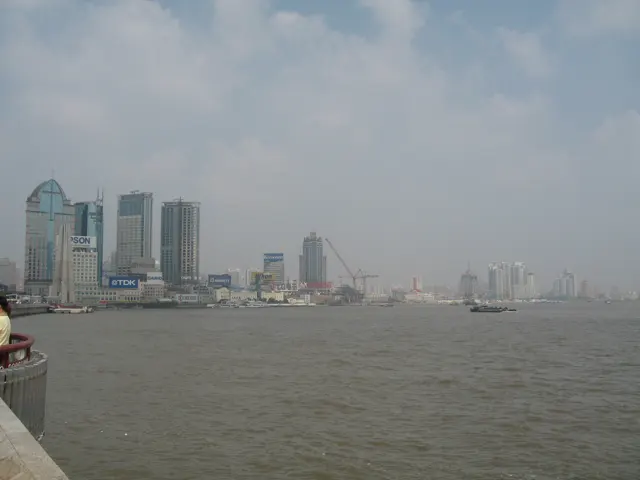 Boats and distant buildings stretch along a broad river beneath a hazy sky.