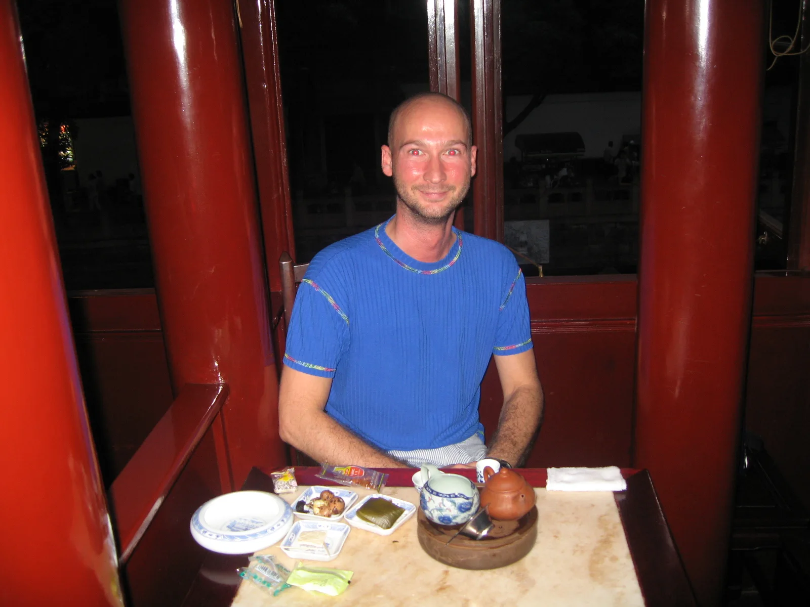 A person in a blue shirt sits at a table with a teapot and cups between red interior columns.