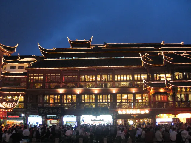 A crowd gathers below a long building whose layered roofs and windows are outlined in warm light.