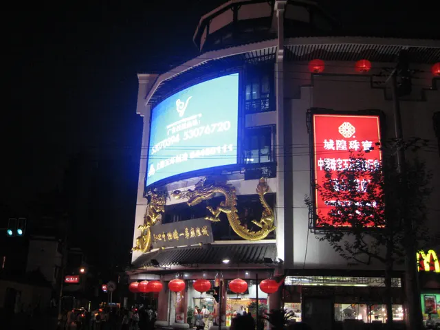 Large illuminated advertising screens rise above a street and red lanterns against the dark sky.