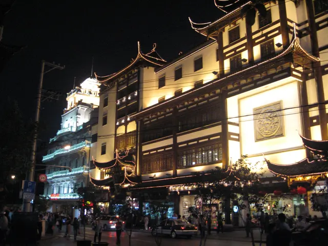 A busy night-time shopping street runs beside brightly lit facades with curved traditional-style roofs.