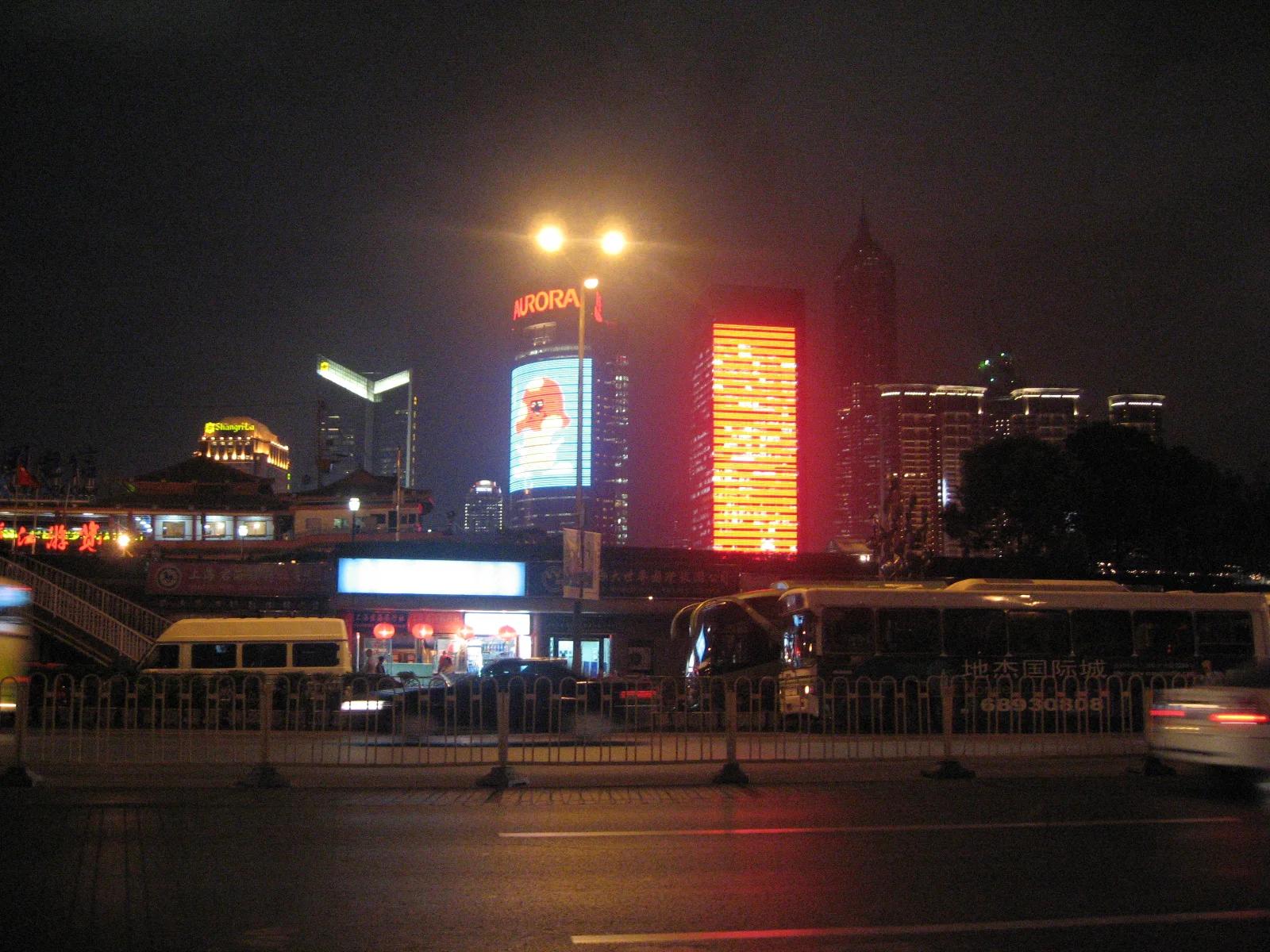 Bright building displays and street lamps illuminate a night-time road and its reflective surface.