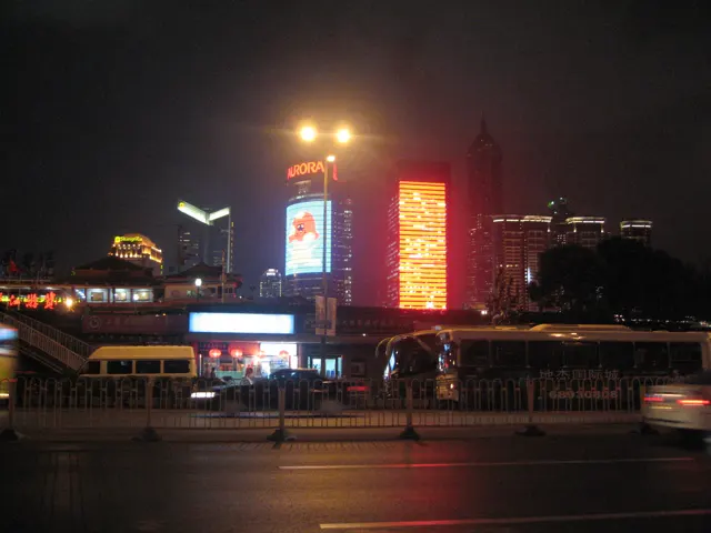 Bright building displays and street lamps illuminate a night-time road and its reflective surface.