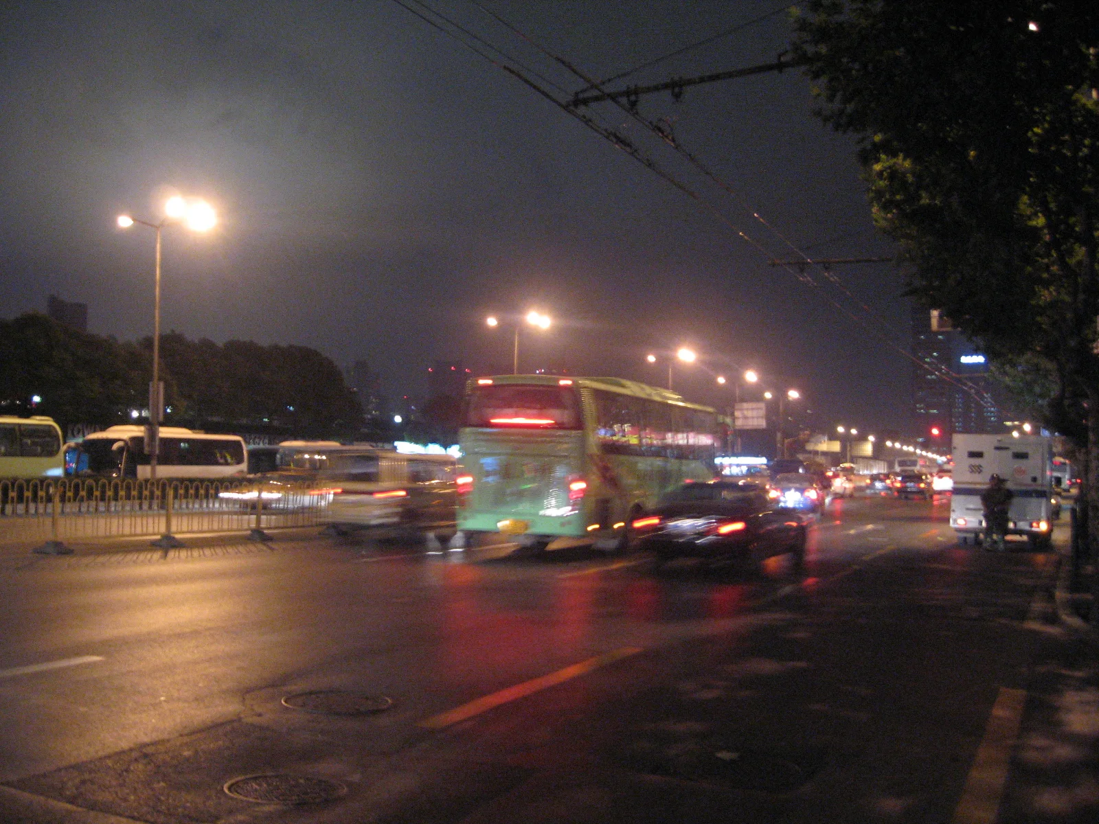 Buses and cars move along a wet road beneath street lamps, leaving coloured reflections across the surface.