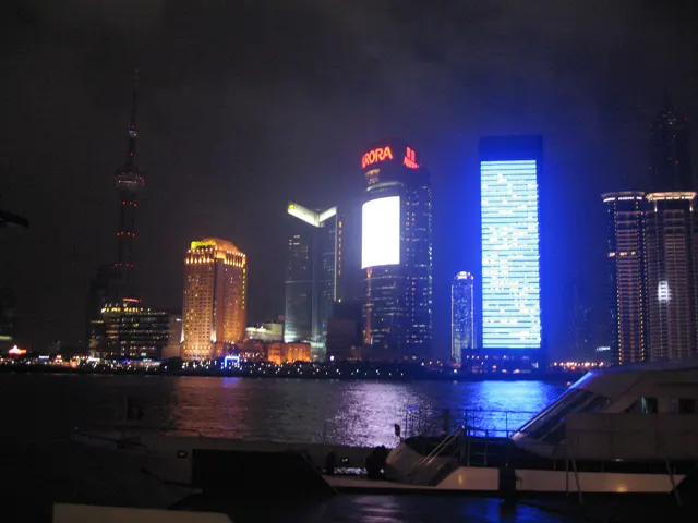 A tall blue-lit building reflects on the dark river beside other illuminated towers.