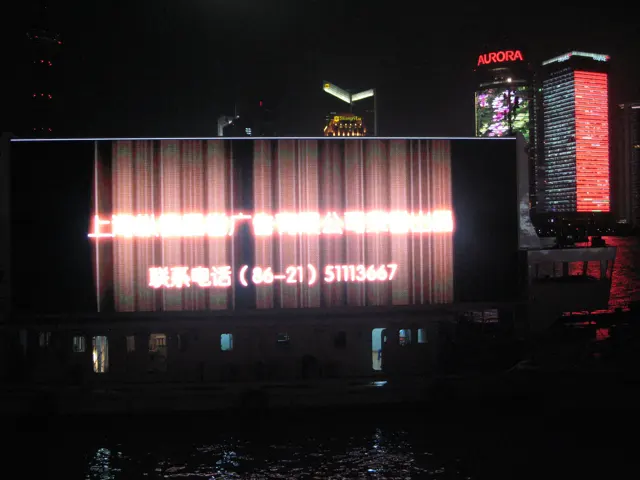 A bright rectangular display shines against dark water, with illuminated buildings behind it.