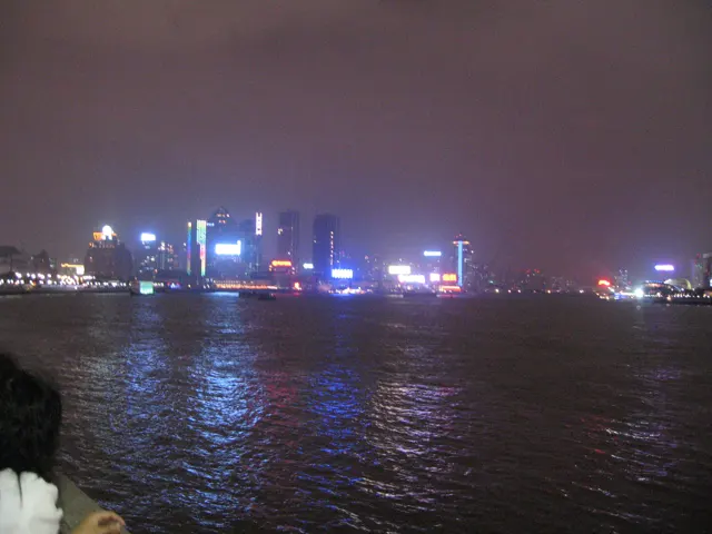 Small blue and white lights line the far shore, reflected in rippling dark water beneath a hazy sky.