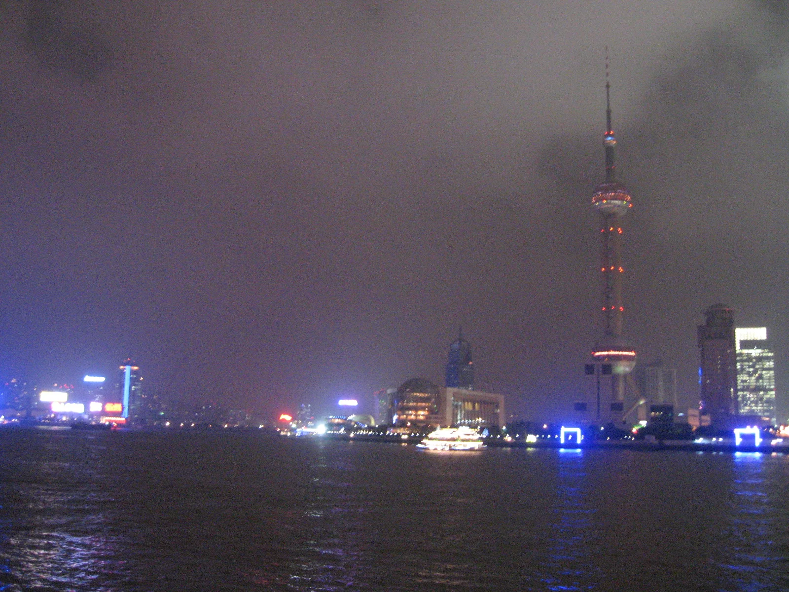 A tall tower with spherical sections rises above the illuminated waterfront beneath low grey clouds.