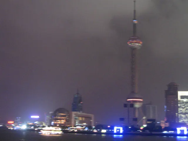A slender tower with a lit spherical section stands above bright blue waterfront lights in a hazy night sky.