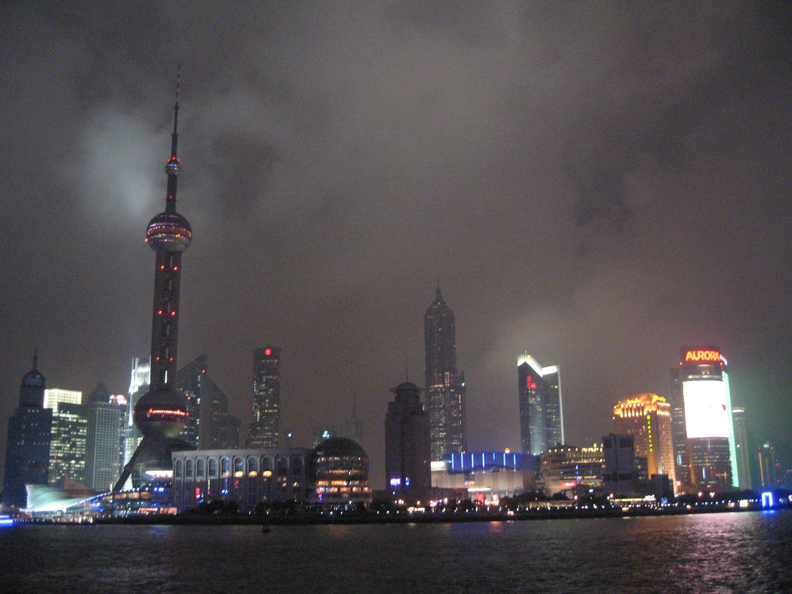 Bright towers and waterfront lights form a skyline across the river beneath a cloud-filled night sky.