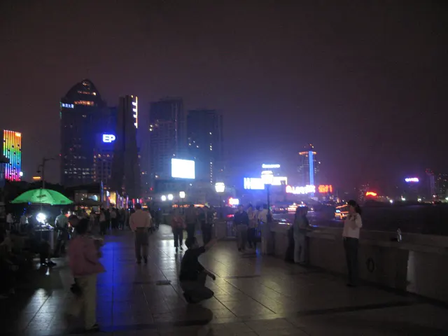 People walk along a wet riverside promenade with bright city lights reflected on the pavement.
