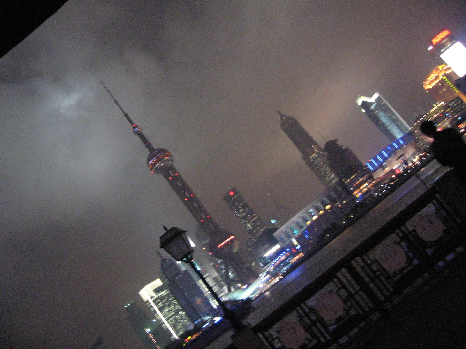 A tilted night view frames the tower and skyline above a dark decorative riverside railing.