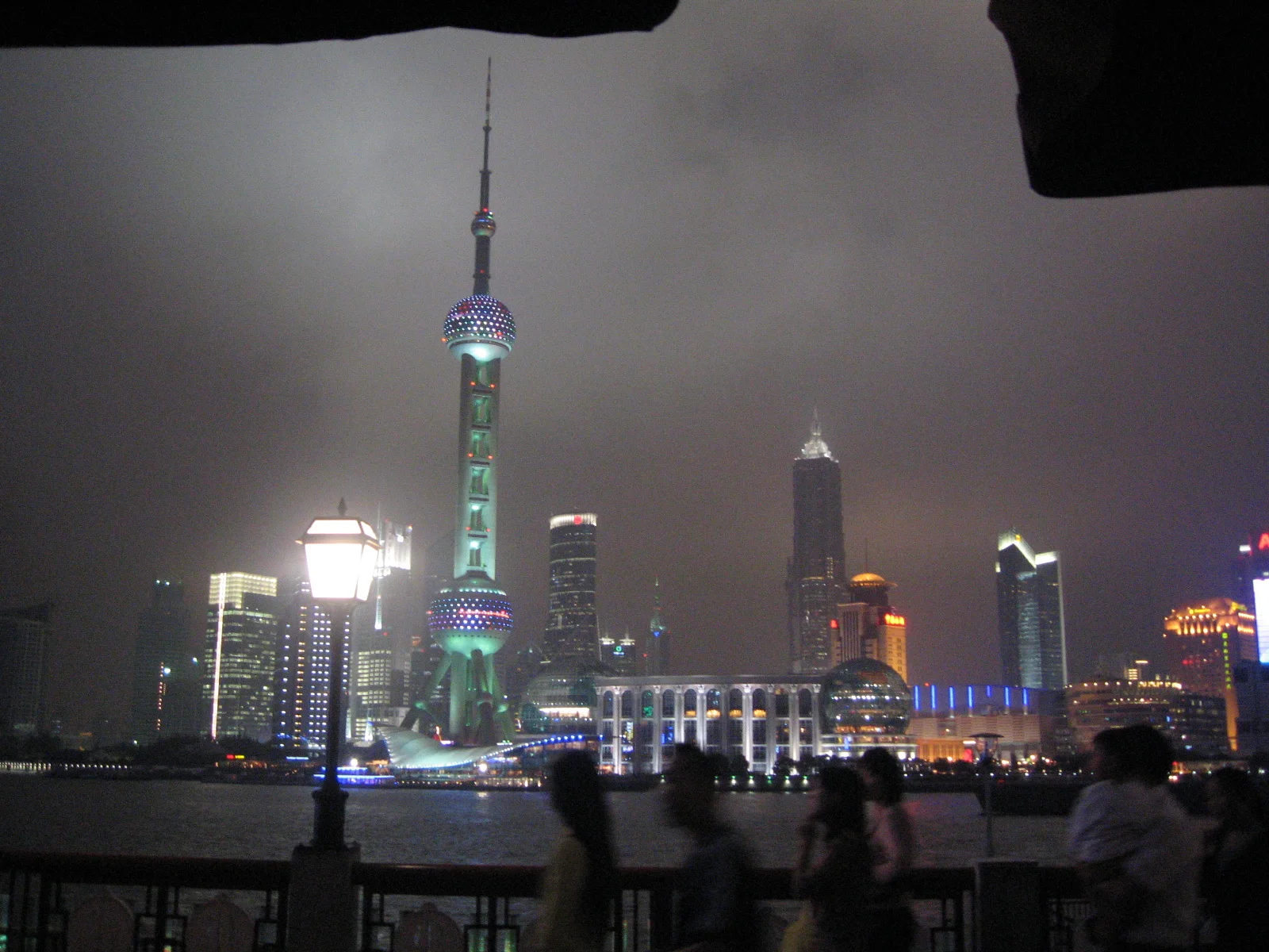 People gather at a riverside railing beneath a skyline dominated by a green-lit tower with spherical sections.