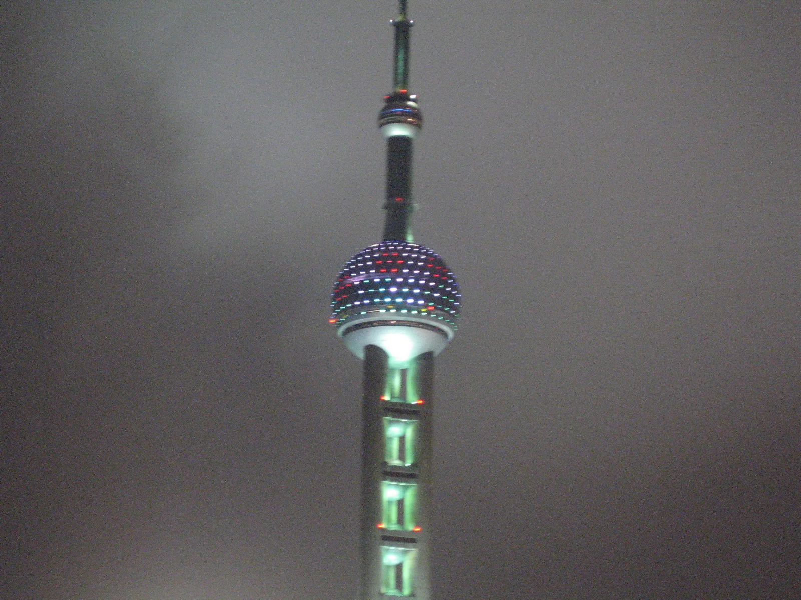 A close night view shows a tower sphere ringed with coloured lights on a green-lit shaft against grey clouds.