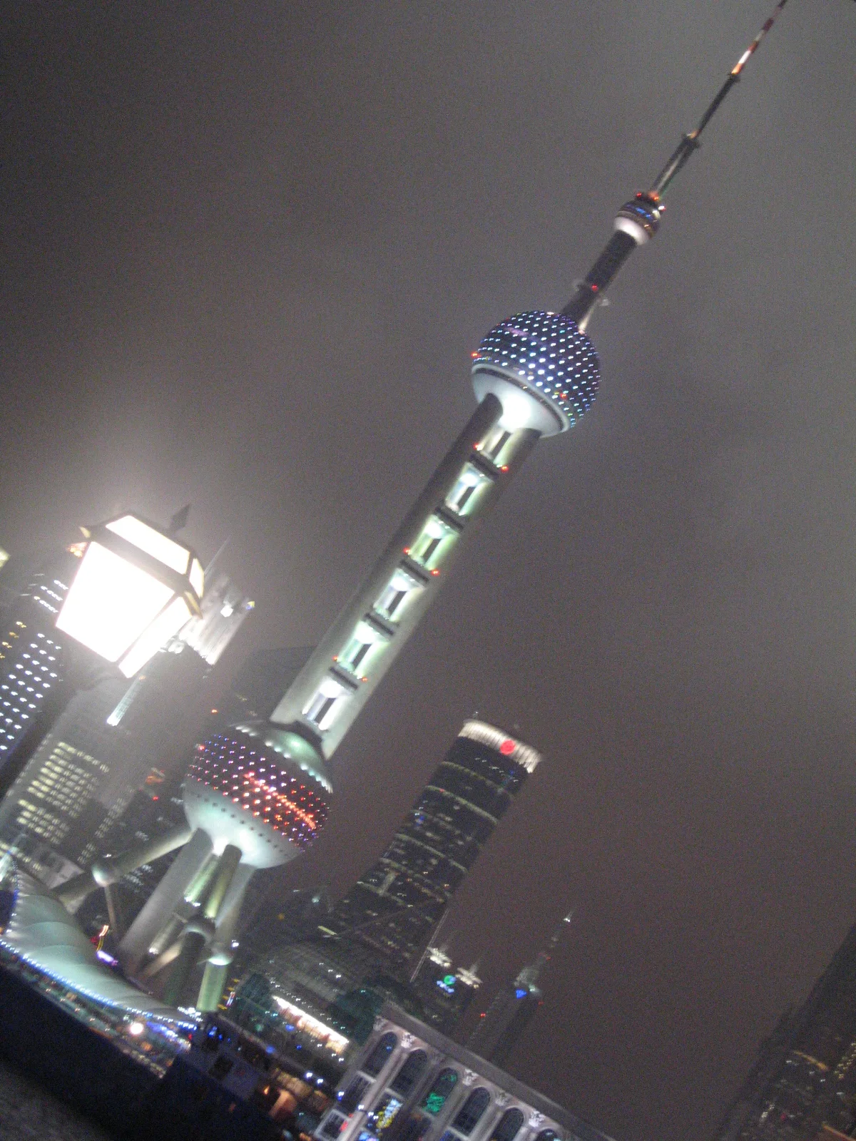 A tilted portrait view follows a brightly lit tower into a cloudy night sky beside a street lamp.