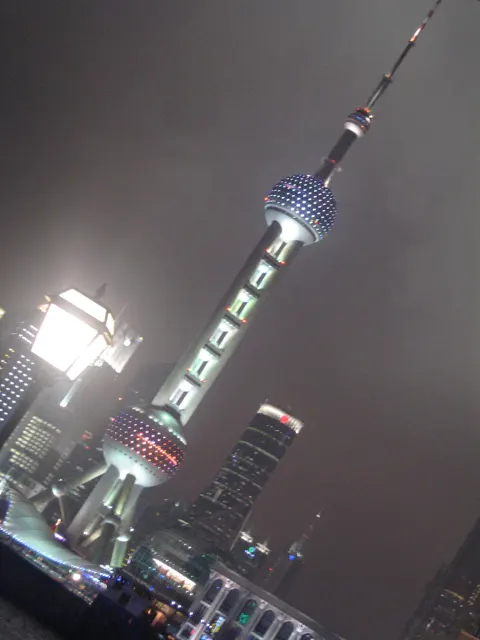 A tilted portrait view follows a brightly lit tower into a cloudy night sky beside a street lamp.
