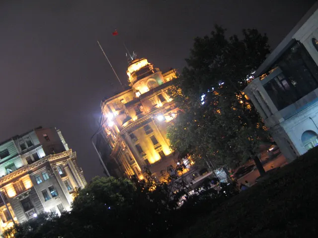 An angled view shows warmly illuminated buildings with towers and ornate details behind dark trees.