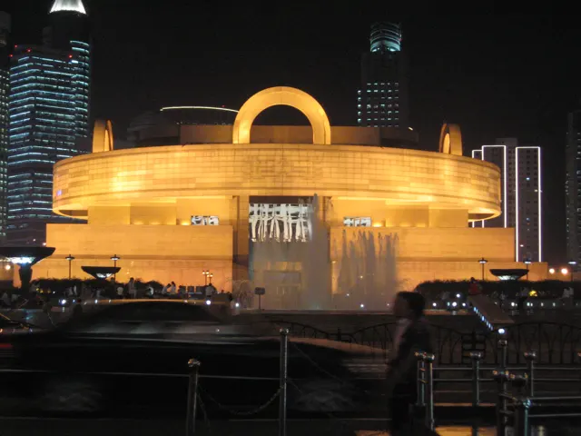 A broad rounded building glows gold at night, with a ring-shaped feature above its centre and taller buildings behind.