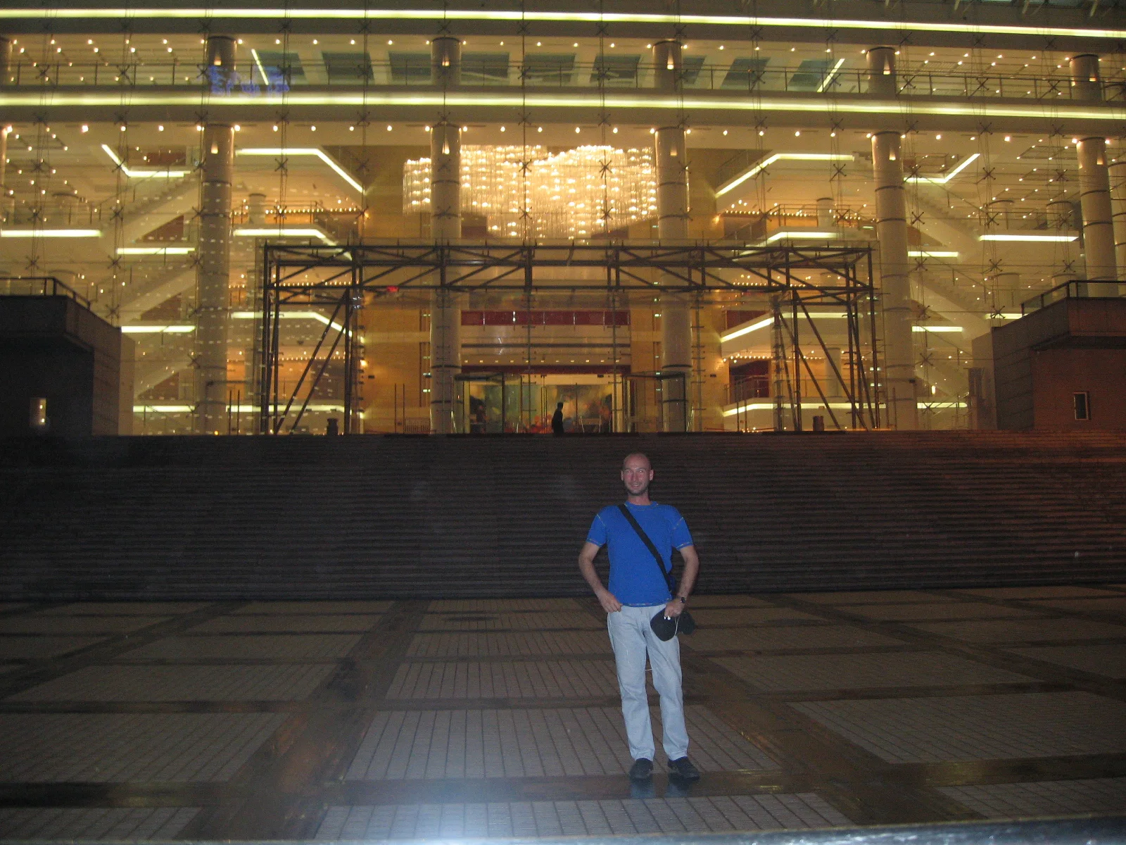 A person in a blue shirt stands on a wide paved forecourt before a brightly lit glass building entrance.