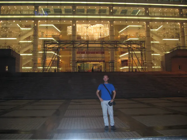 A person in a blue shirt stands on a wide paved forecourt before a brightly lit glass building entrance.