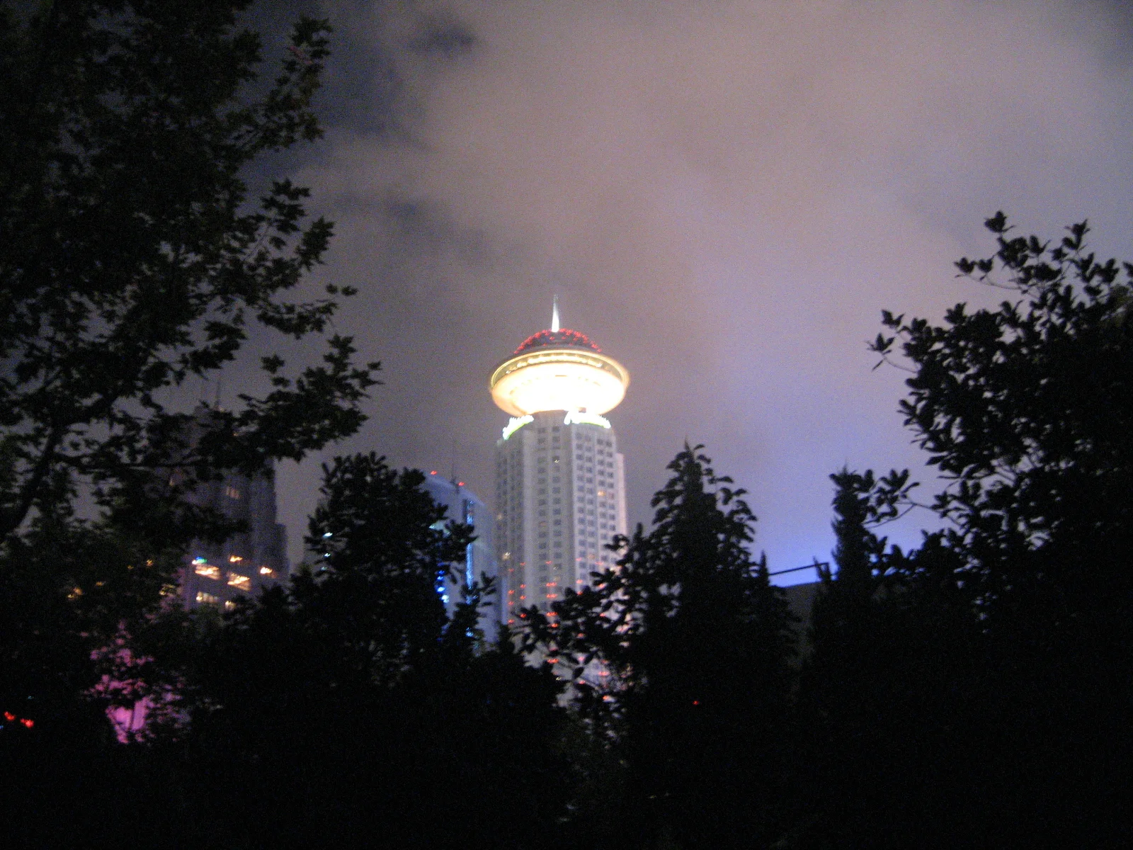 A tower with a glowing circular top rises behind silhouetted trees in a purple-grey night sky.