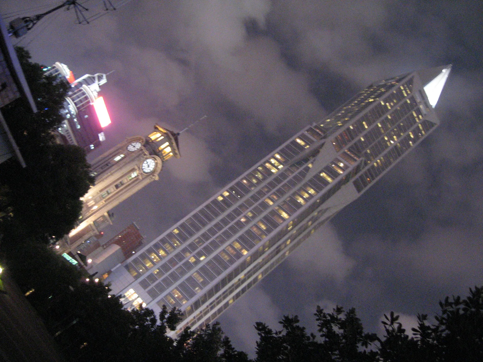 A tilted upward night view follows a tall illuminated building with a pointed crown beside dark foliage.
