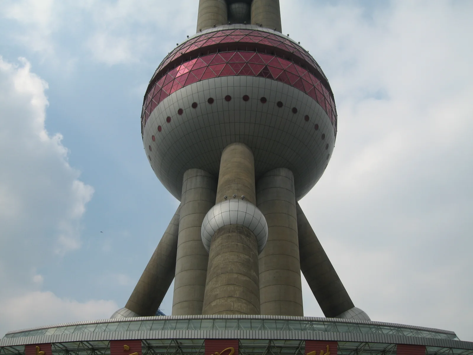 A broad pink-banded sphere rests on large sloping supports above an entrance structure beneath clouds.