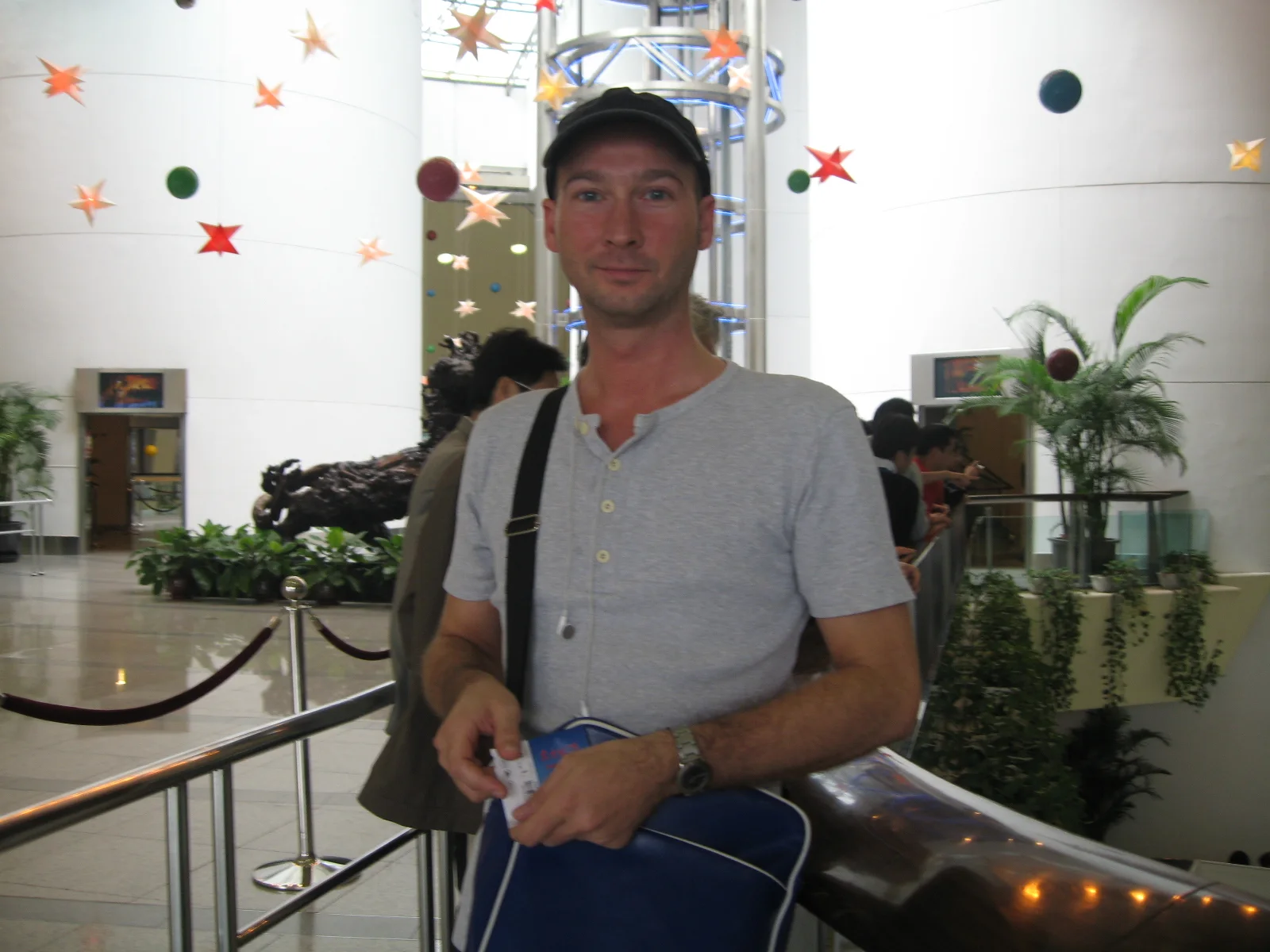 A person wearing a cap and shoulder bag stands by an indoor railing, with plants and hanging decorations behind.