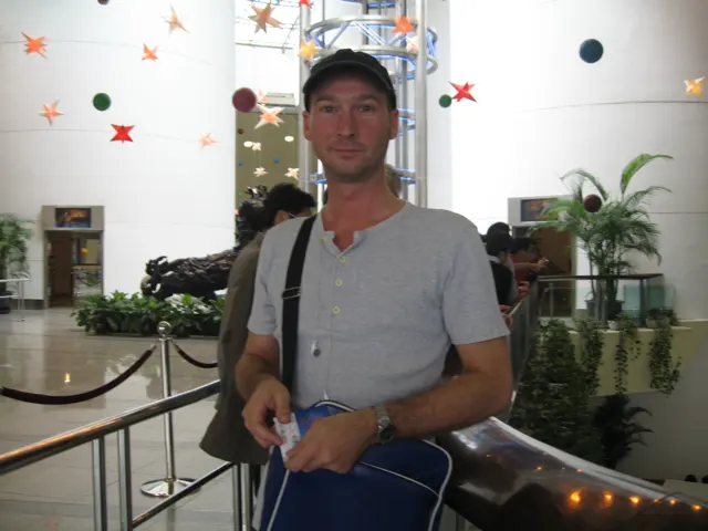 A person wearing a cap and shoulder bag stands by an indoor railing, with plants and hanging decorations behind.