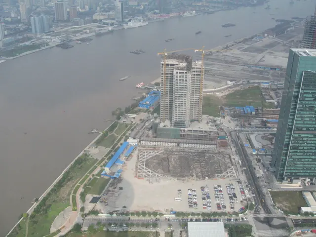 An elevated view looks down over a broad river, waterfront buildings and a large paved area beside a tall glass facade.