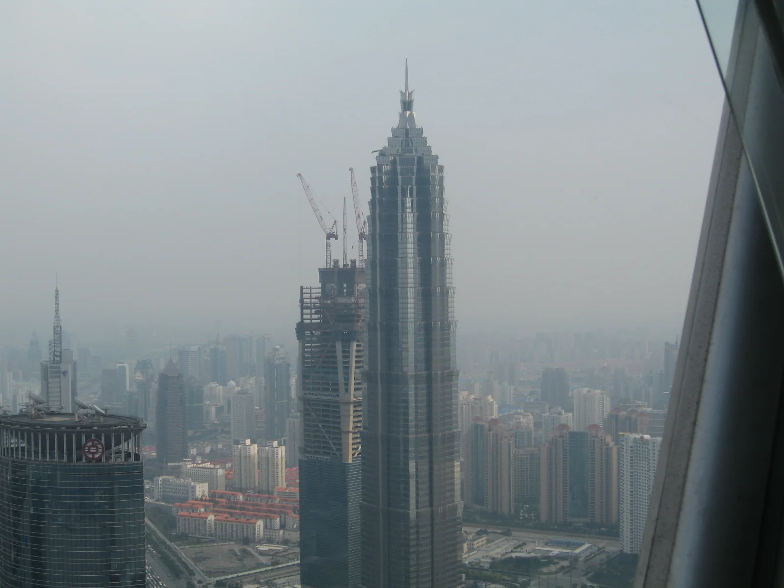 A tall stepped skyscraper and nearby construction appear through haze beyond an angled window frame.