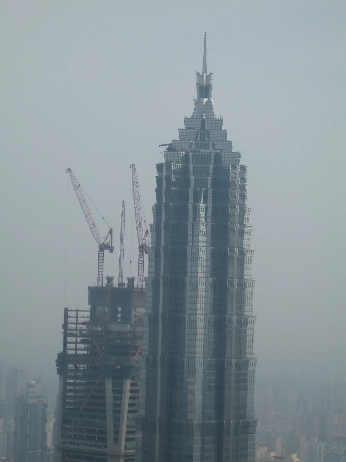 A closer elevated view shows a pointed high-rise beside a construction crane against a pale hazy skyline.