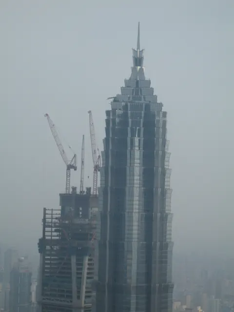 A closer elevated view shows a pointed high-rise beside a construction crane against a pale hazy skyline.